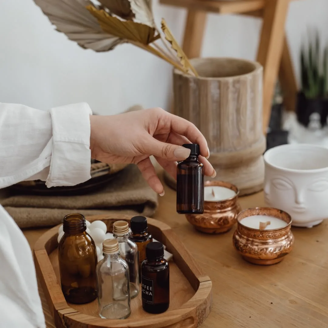 A hand holding a small amber bottle of fragrance oil above a tray of perfume-making bottles and copper candles